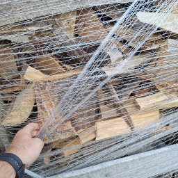 PRYMEK Arkadiusz Kołakowski - Ręka w czarnym zegarku dotyka spakowanego w folię pakietu pociętego drewna na palecie.