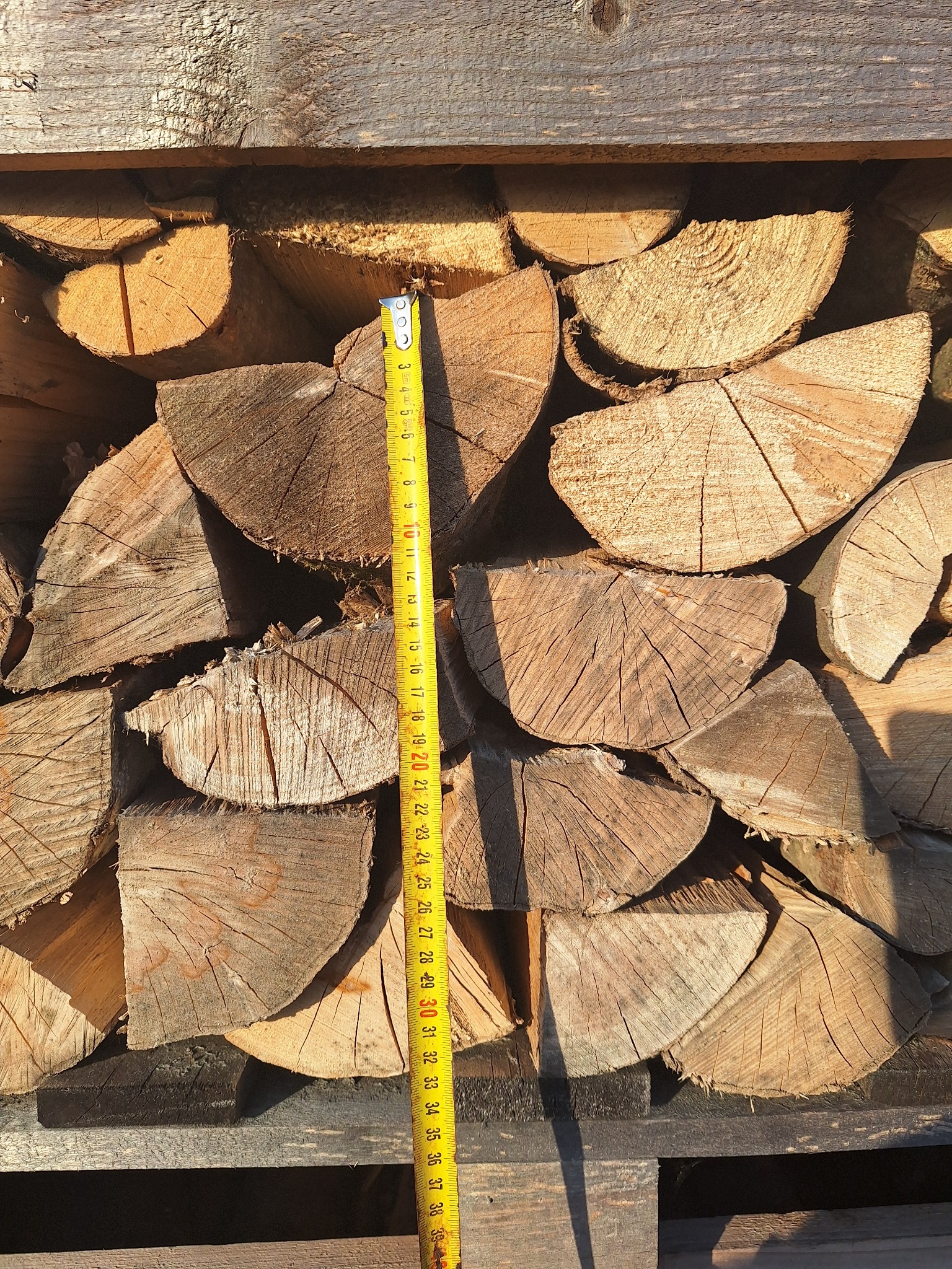 Ułożone, porąbane kawałki drewna na palecie, z żółtą miarką pionowo pośrodku, wskazującą na ich przybliżoną długość.