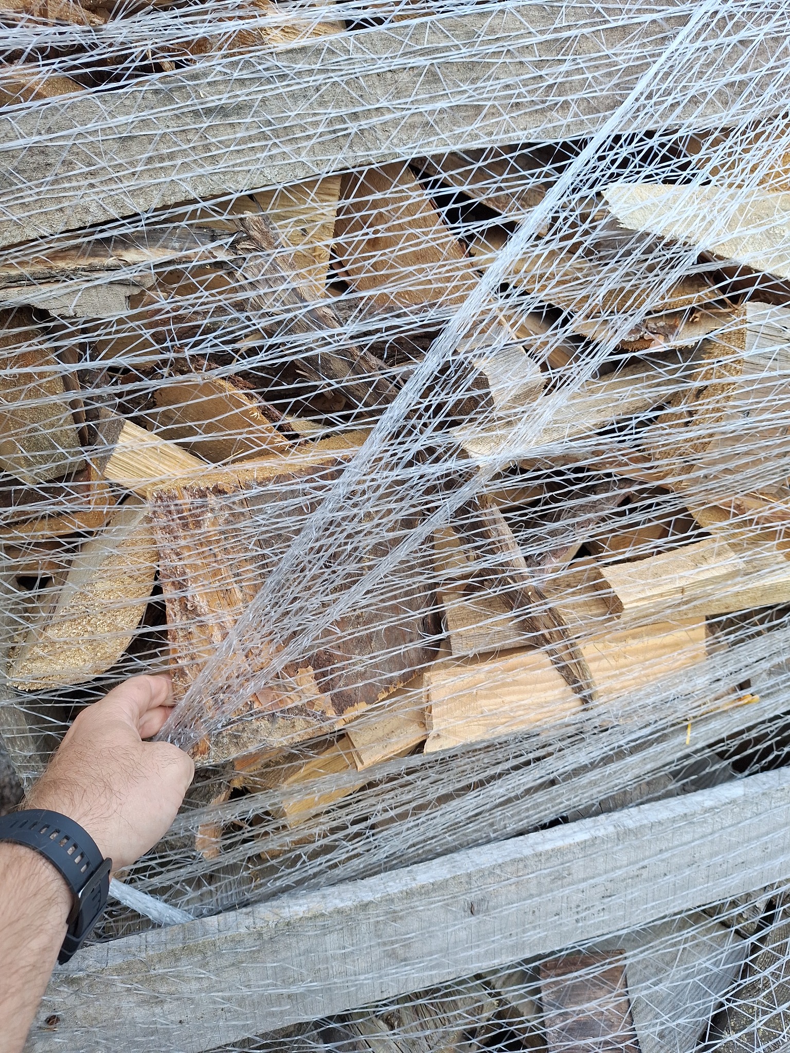 Ręka w czarnym zegarku dotyka spakowanego w folię pakietu pociętego drewna na palecie.