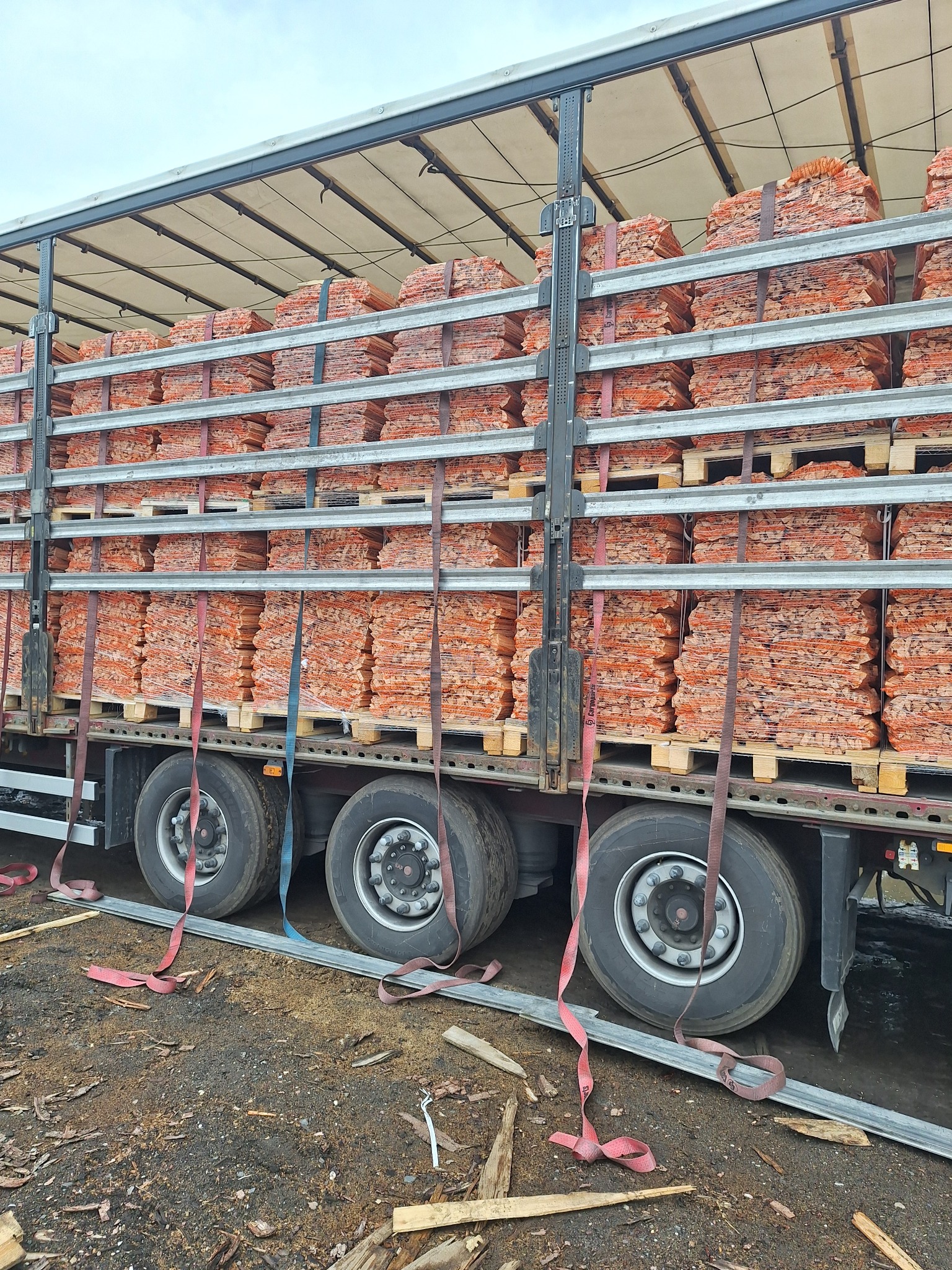 Ciężarówka załadowana paletami z drewnem opałowym, zabezpieczonymi pasami, widoczna z boku na placu składowym z fragmentami drewna na ziemi.