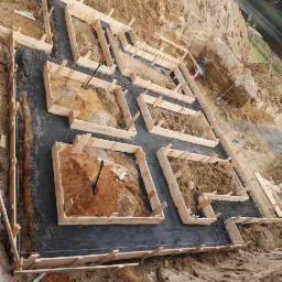 Zalane fundamenty pod budynek z drewnianym szalunkiem i wystającą stalową siatką zbrojeniową, widok z góry na wykopie w ziemi.