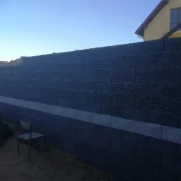 Wznoszenie muru oporowego z ciemnych bloczków betonowych, widoczna warstwa jasnych elementów poziomujących, krzesło robocze w pobliżu, w tle budynek mieszkalny.