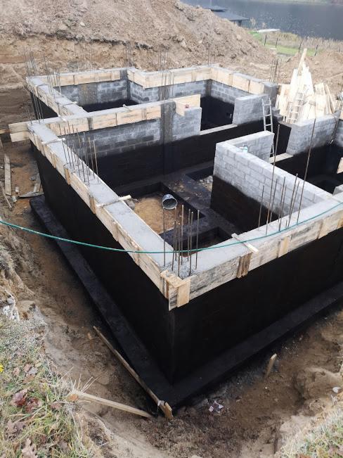 Budowa fundamentów domu murowanego: wylany beton, zbrojenie wystające pionowo, izolacja przeciwwilgociowa na ścianach fundamentowych, szalunki drewniane, widoczne wykopy ziemne.