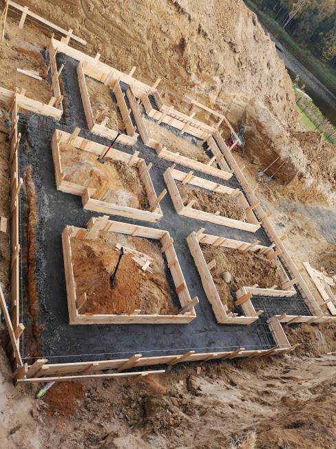 Zalane fundamenty pod budynek z drewnianym szalunkiem i wystającą stalową siatką zbrojeniową, widok z góry na wykopie w ziemi.