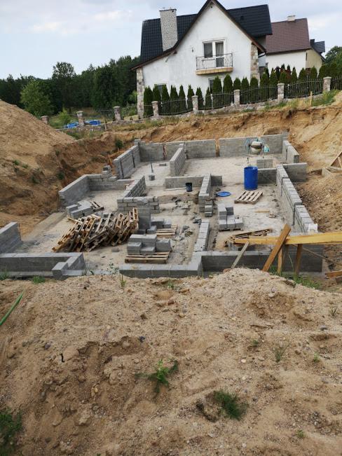 Wykopane fundamenty pod budynek w trakcie wznoszenia, widoczne bloczki betonowe, palety i niebieski pojemnik na wodę, w tle dom jednorodzinny z balkonem.