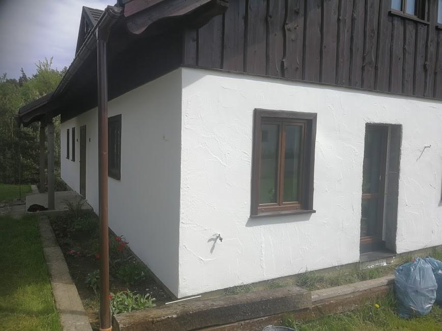 Świeżo otynkowany, biały dom z drewnianą podbitką dachową i brązowymi ramami okiennymi. Widoczna elewacja z fakturą baranka, rynna spustowa i fragment ogrodu z kwiatami.