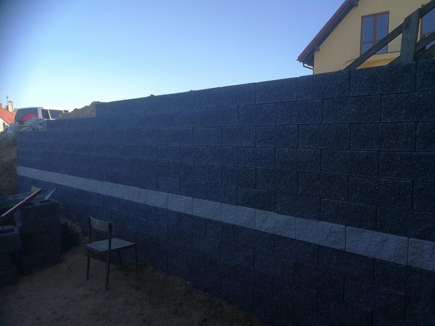 Wznoszenie muru oporowego z ciemnych bloczków betonowych, widoczna warstwa jasnych elementów poziomujących, krzesło robocze w pobliżu, w tle budynek mieszkalny.