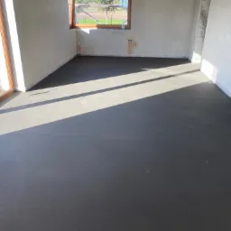 Świeżo wylana, gładka, ciemnoszara posadzka jastrychowa w jasnym pomieszczeniu z oknami i wstępnym wykończeniem ścian.