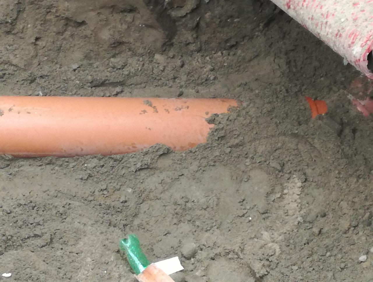 Pomarańczowa rura kanalizacyjna ułożona w wykopie ziemnym, widoczne fragmenty innych rur i narzędzi.