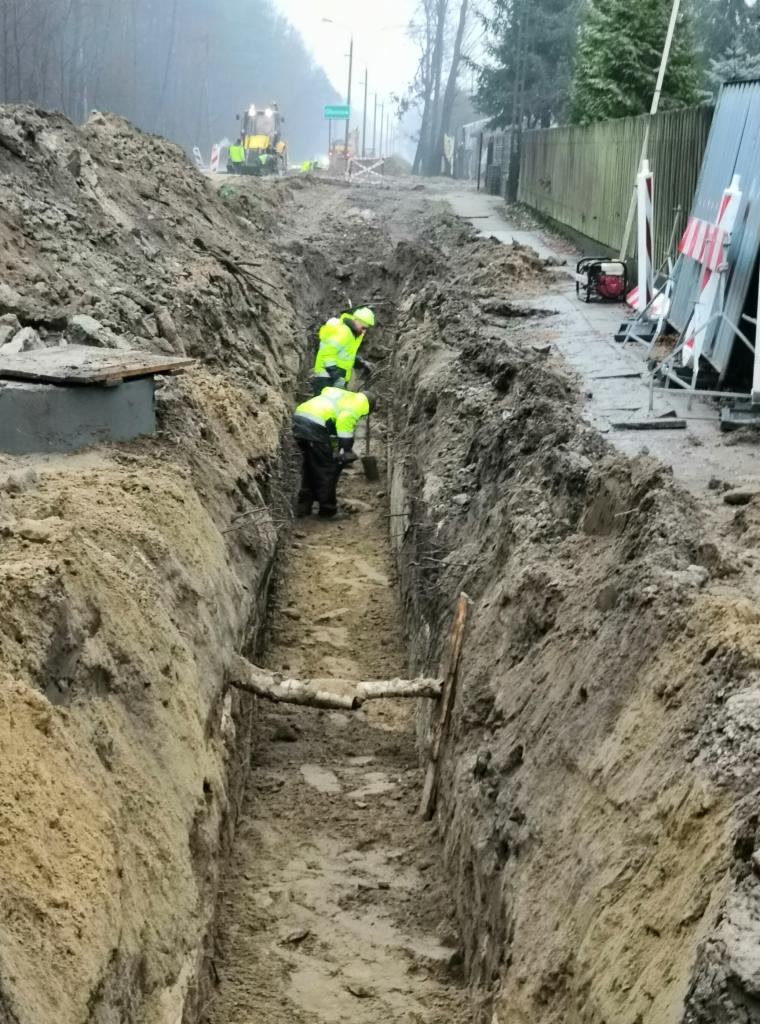 Głęboki wykop w ziemi z dwoma pracownikami w odblaskowych kamizelkach, pracującymi łopatami, z widoczną podporą z gałęzi, na tle maszyna budowlana i miejski krajobraz w pochmurny dzień.