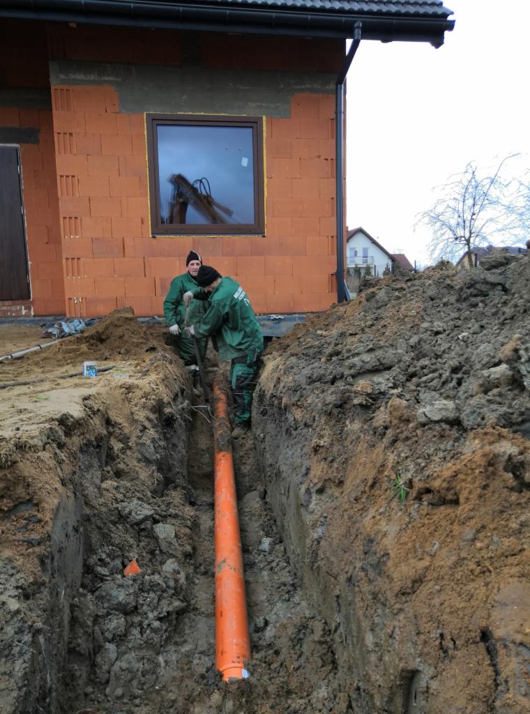 Dwaj pracownicy w zielonych kombinezonach umieszczają pomarańczową rurę w wykopie obok budynku w trakcie budowy. W tle widoczny krajobraz z domami.