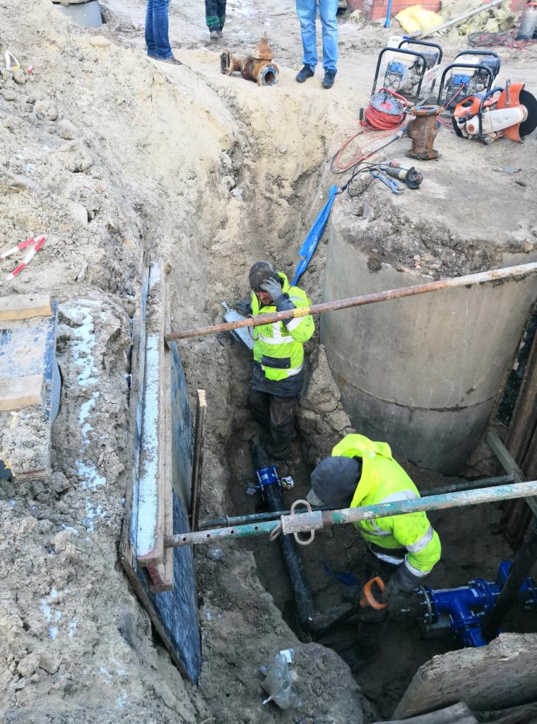 Pracownicy w wykopie montują rury wodociągowe; widoczne szalunki, betonowy krąg i narzędzia na powierzchni gruntu.
