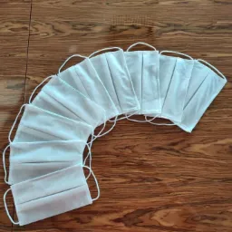 Rząd białych, plisowanych maseczek ochronnych z gumkami, ułożonych wachlarzowo na ciemnym drewnianym blacie.