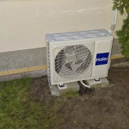 Zewnętrzna jednostka klimatyzatora Haier zamontowana na betonowych bloczkach przy ścianie domu, widoczna trawa i krzew.