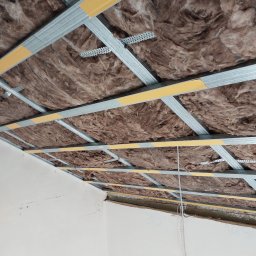 Tobiasz Chamioło INTER-TECH - Montaż izolacji z wełny mineralnej i metalowej konstrukcji sufitu podwieszanego. Widoczne profile, taśmy uszczelniające i fragment ściany z oznaczeniami.