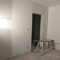 Tobiasz Chamioło INTER-TECH - Pomieszczenie w trakcie prac tynkarskich, z częściowo nałożoną, fakturowaną warstwą na ścianie, widocznym otworem drzwiowym, betonową podłogą z czerwonymi liniami i narzędziami budowlanymi.