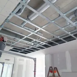 Montaż metalowego stelażu sufitu podwieszanego z widocznymi kanałami wentylacyjnymi i fragmentem płyty kartonowo-gipsowej, drabina w tle.