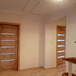 Jasne wnętrze z drewnianą podłogą i dwiema drewnianymi drzwiami z poziomymi przeszkleniami, widoczny fragment ozdobnego żyrandola i klapa rewizyjna w suficie.