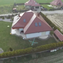 Widok z lotu ptaka na dom jednorodzinny z czerwoną dachówką, otoczony zielonym żywopłotem i trawnikiem z ozdobnymi nasadzeniami, w tle pola uprawne i inne domy.