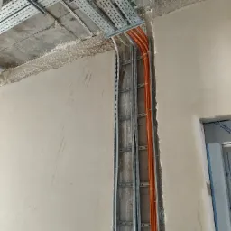 Montaż korytek kablowych i przewodów elektrycznych w surowym stanie budynku, widoczne pomarańczowe i białe kable prowadzone w metalowych korytach, surowy tynk na ścianach i suficie.