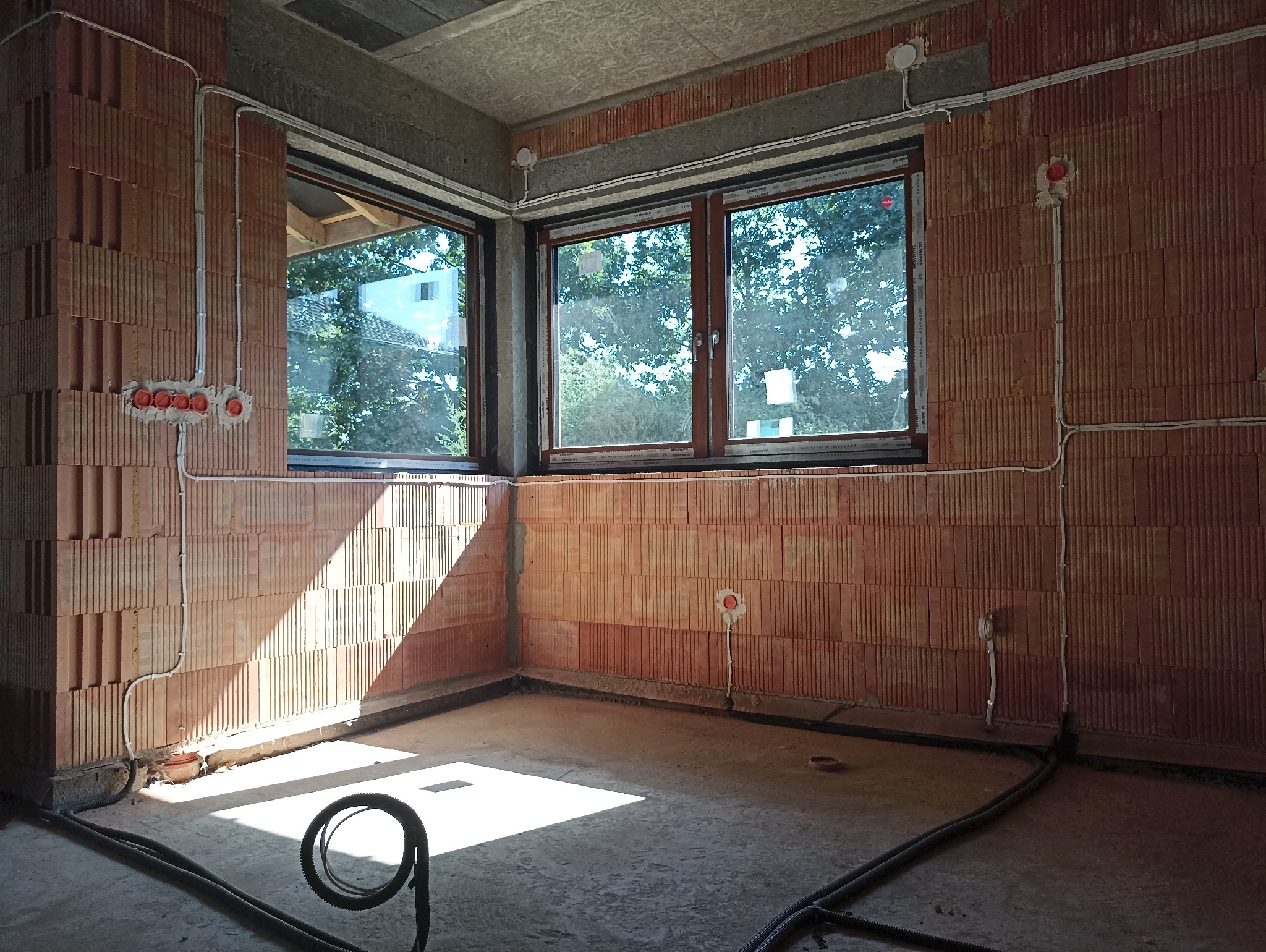 Surowy mur z czerwonej cegły dziurawki z widocznymi puszkami instalacyjnymi i przewodami elektrycznymi, dwa okna z widokiem na zieleń, wpadające światło słoneczne na betonową podłogę w trakcie prac...