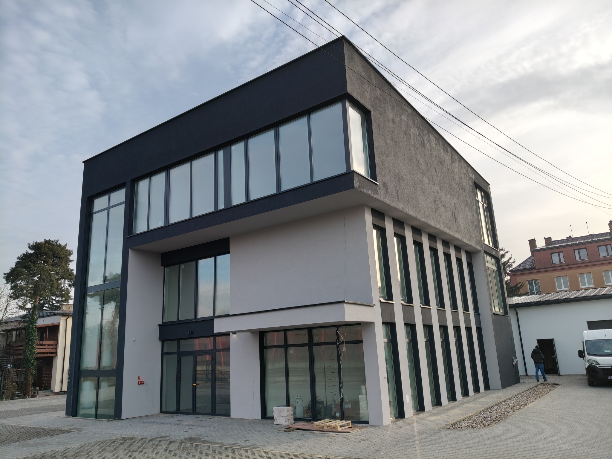 Nowoczesny budynek biurowy z szarymi i czarnymi elewacjami, duże okna, widoczne przewody elektryczne na zewnątrz budynku, kostka brukowa na podjeździe.