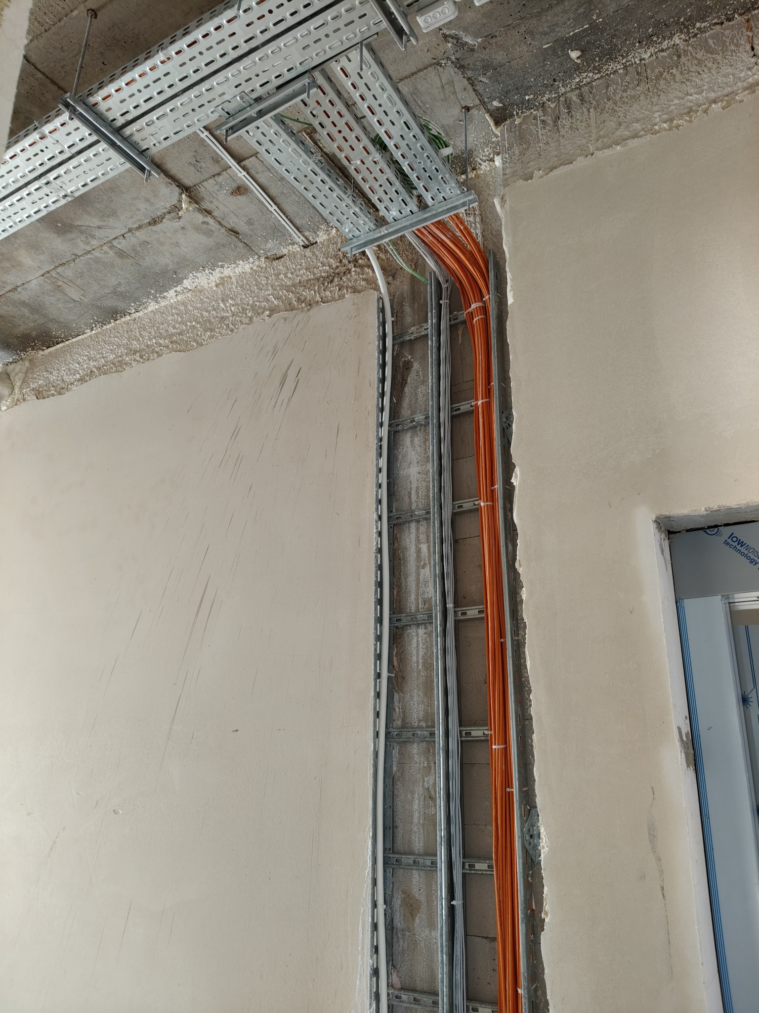 Montaż korytek kablowych i przewodów elektrycznych w surowym stanie budynku, widoczne pomarańczowe i białe kable prowadzone w metalowych korytach, surowy tynk na ścianach i suficie.