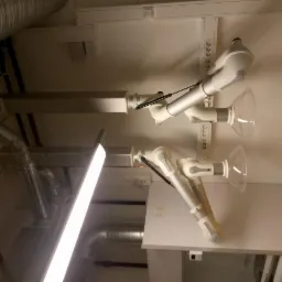 Białe, regulowane ramiona odciągowe z przezroczystymi okapami, zamontowane na ścianie w pomieszczeniu z widocznymi rurami wentylacyjnymi pod sufitem.