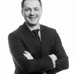 Portret uśmiechniętego mężczyzny w ciemnym garniturze, z rękami skrzyżowanymi na piersiach, na jasnym tle.