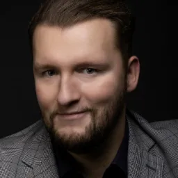 Portret mężczyzny z brodą w szarej marynarce w kratę na ciemnym tle.