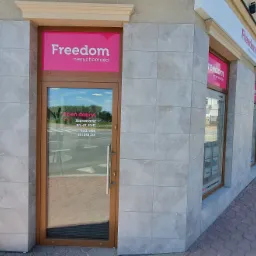 Fasada biura nieruchomości z logo Freedom nieruchomości, wejście z przeszklonymi drzwiami, widoczne godziny otwarcia na szybie, chodnik z kostki brukowej.