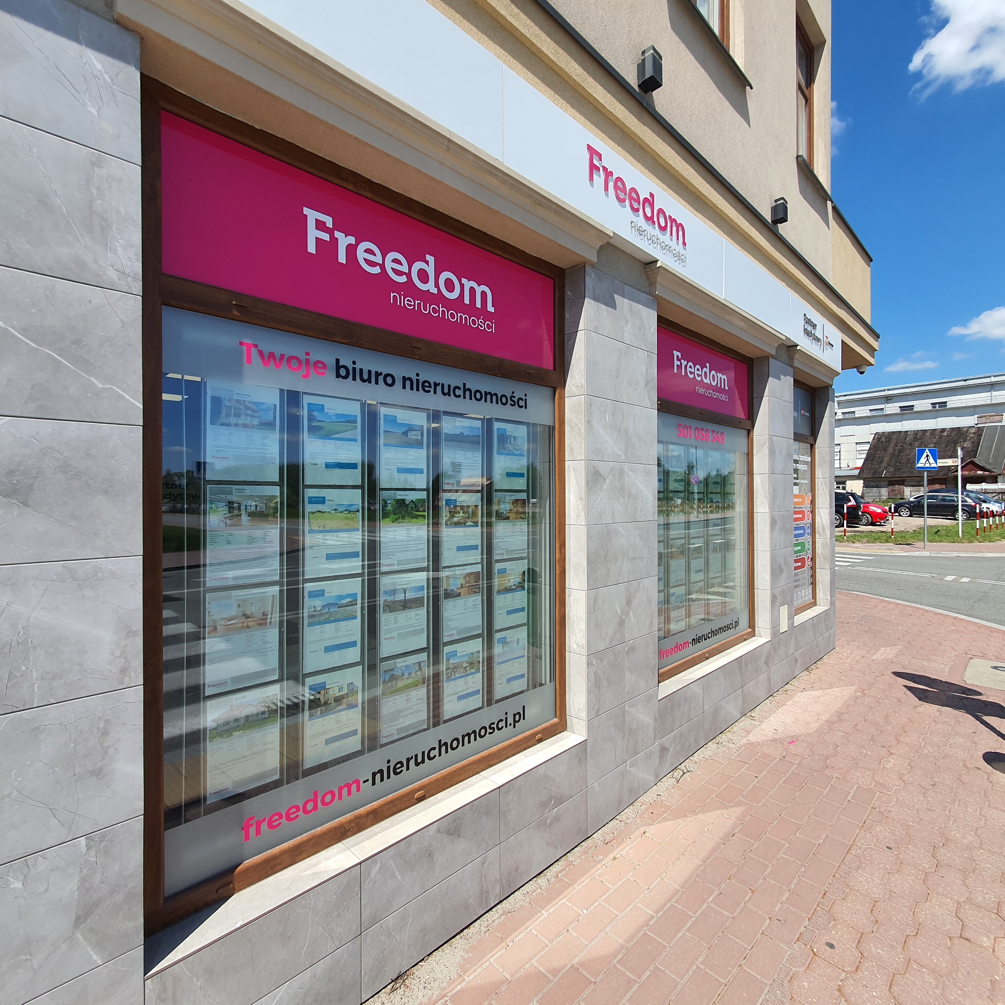 Fasada biura nieruchomości Freedom z witrynami wystawowymi prezentującymi oferty sprzedaży i wynajmu. Na witrynach plakaty z fotografiami nieruchomości. Nad witrynami szyldy z nazwą firmy w kolorze...