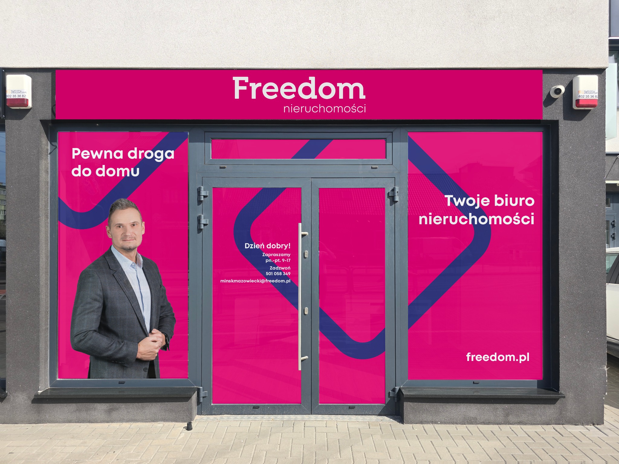 Fasada biura nieruchomości Freedom z dużym logotypem i hasłami reklamowymi na różowym tle, widoczny mężczyzna w garniturze w oknie.