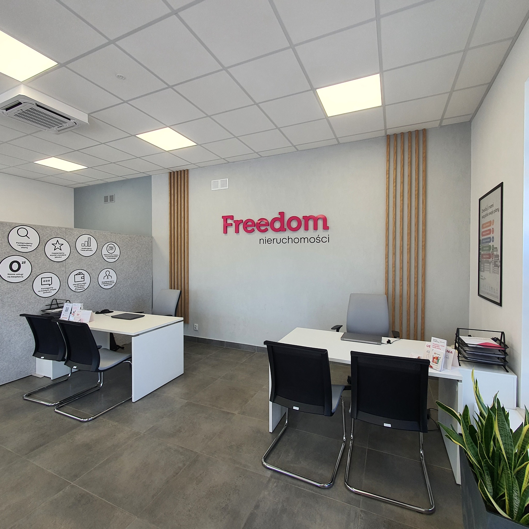 Jasne, nowoczesne biuro nieruchomości z dwoma stanowiskami pracy, logo 'Freedom nieruchomości' na ścianie, rośliną doniczkową i ścianą z informacjami dla klientów.