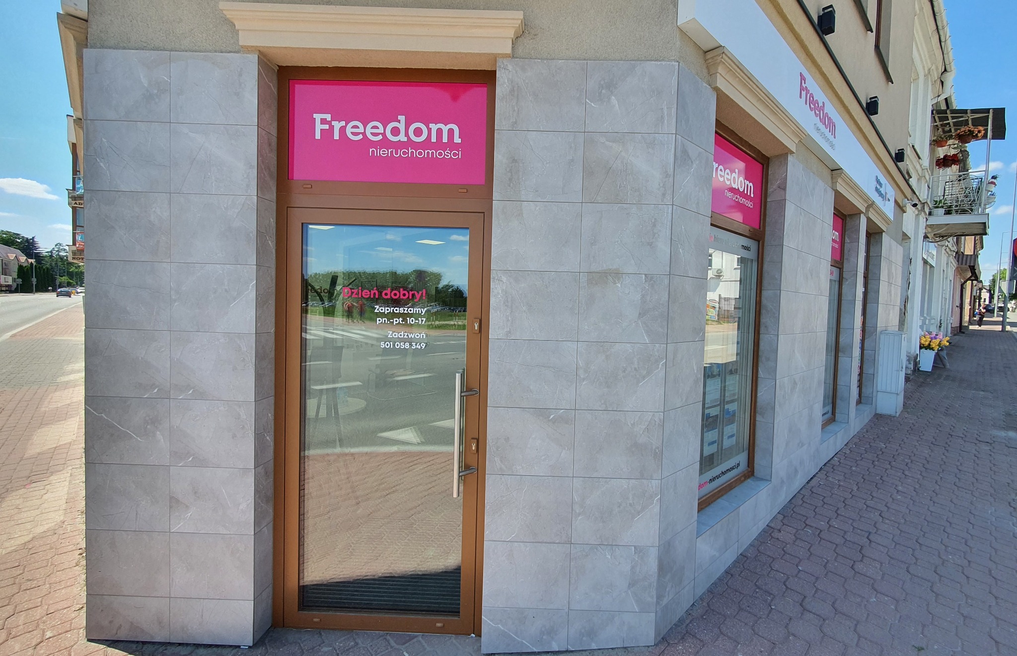 Fasada biura nieruchomości z logo Freedom nieruchomości, wejście z przeszklonymi drzwiami, widoczne godziny otwarcia na szybie, chodnik z kostki brukowej.