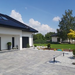 Tevip Investments - Nowoczesny podjazd z szarej kostki brukowej, oświetlony lampami. Biały dom z czarnym dachem i oknami dachowymi w tle. Elementy dekoracyjne z kamienia i roślin.