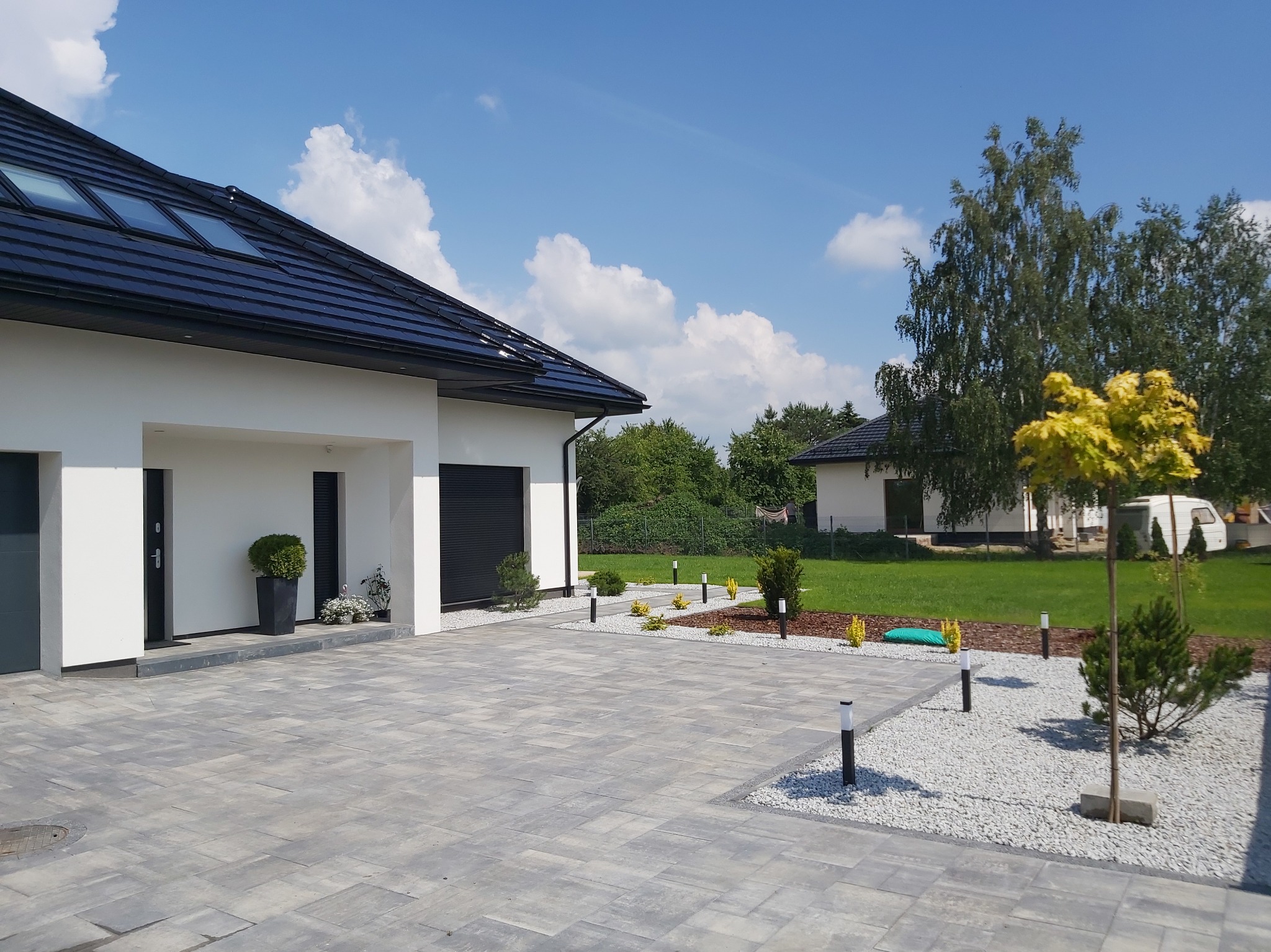 Nowoczesny podjazd z szarej kostki brukowej, oświetlony lampami. Biały dom z czarnym dachem i oknami dachowymi w tle. Elementy dekoracyjne z kamienia i roślin.