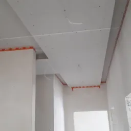 Montaż konstrukcji sufitu podwieszanego z widocznymi profilami i elementami mocującymi na ścianach w surowym wnętrzu.