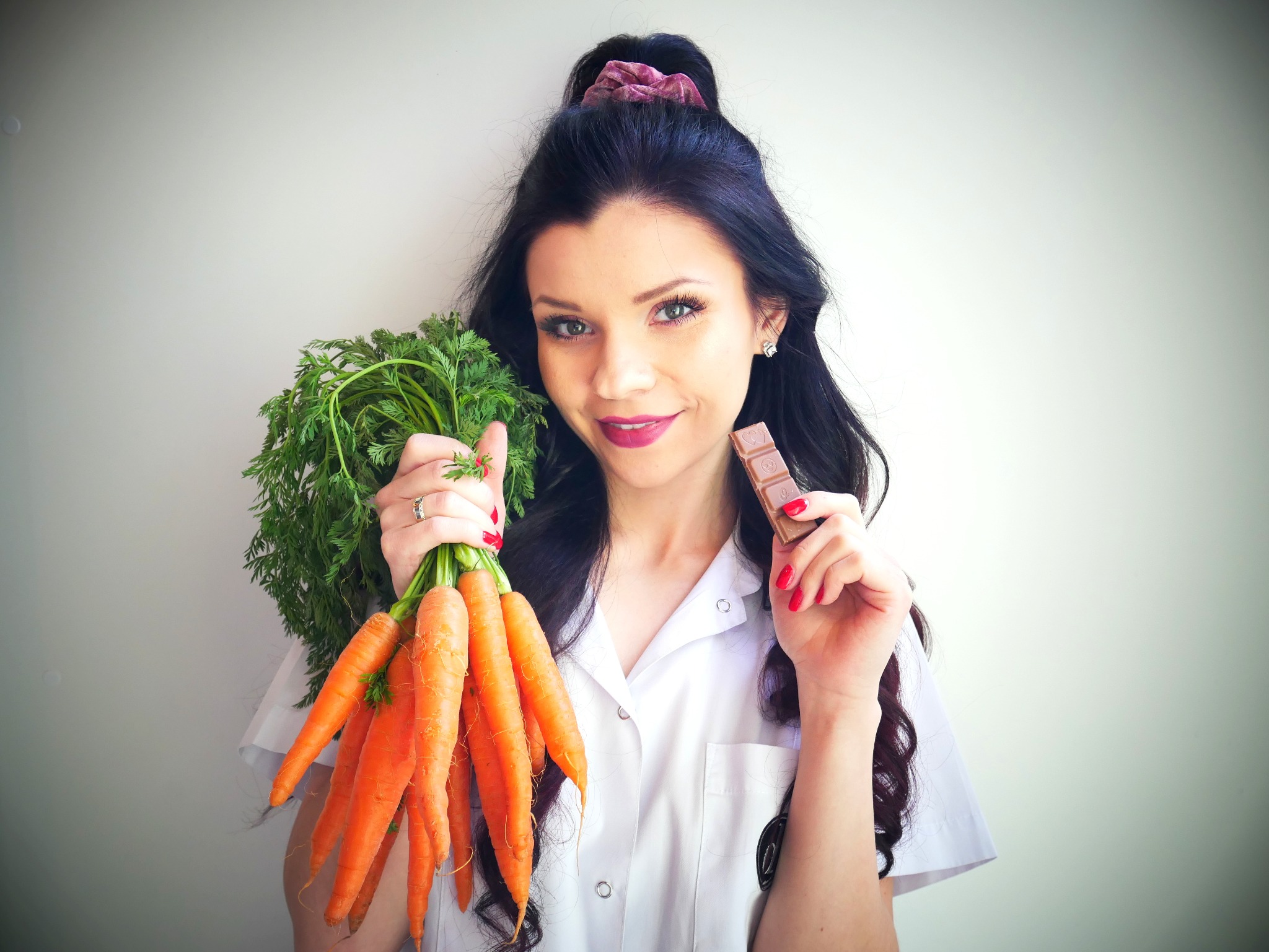 Uśmiechnięta kobieta w białym fartuchu trzyma pęczek marchewek w jednej ręce i czekoladę w drugiej.