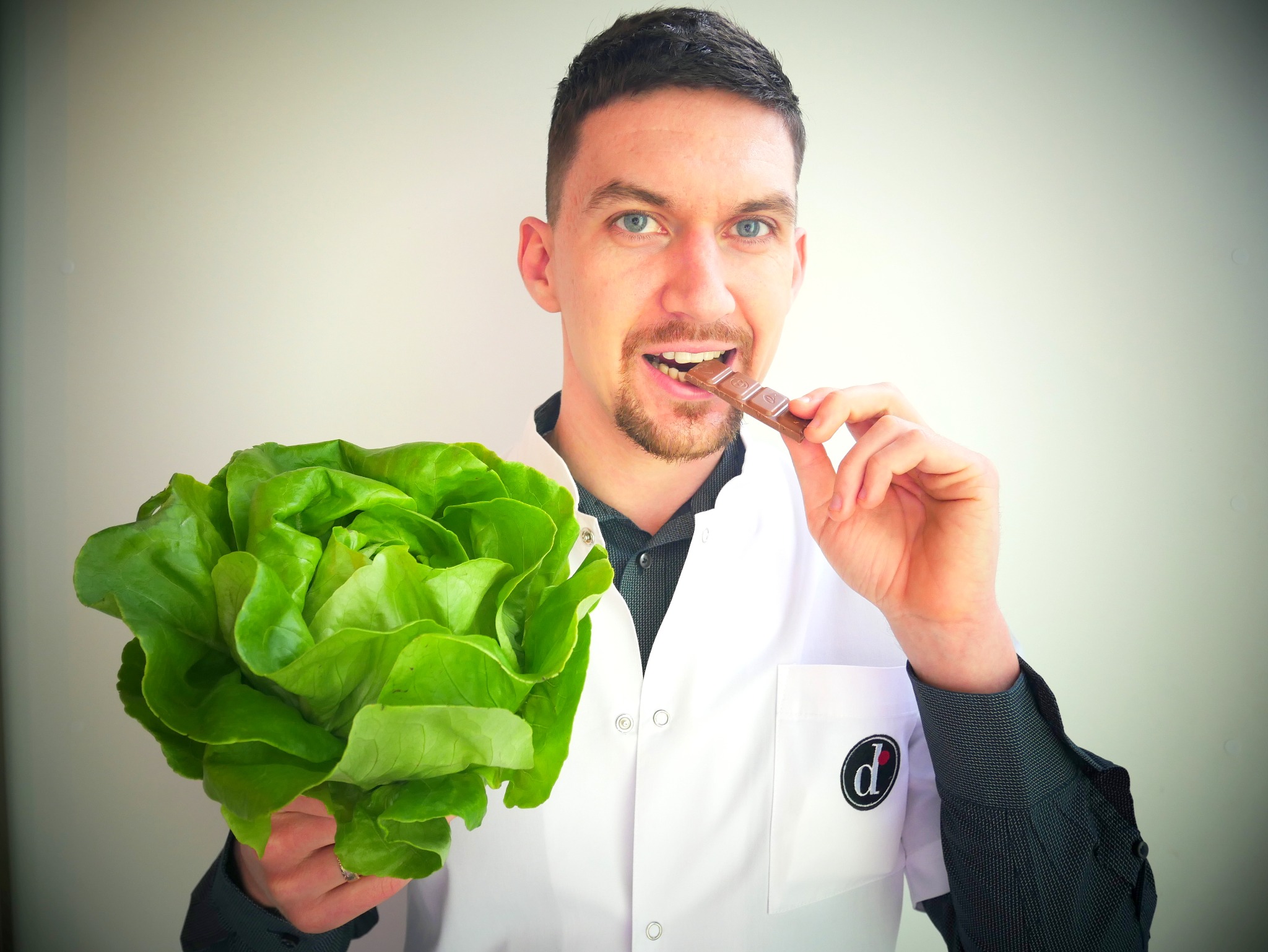 Mężczyzna w białym fartuchu trzyma w jednej ręce główkę sałaty, a w drugiej kostkę czekolady, którą wkłada do ust.