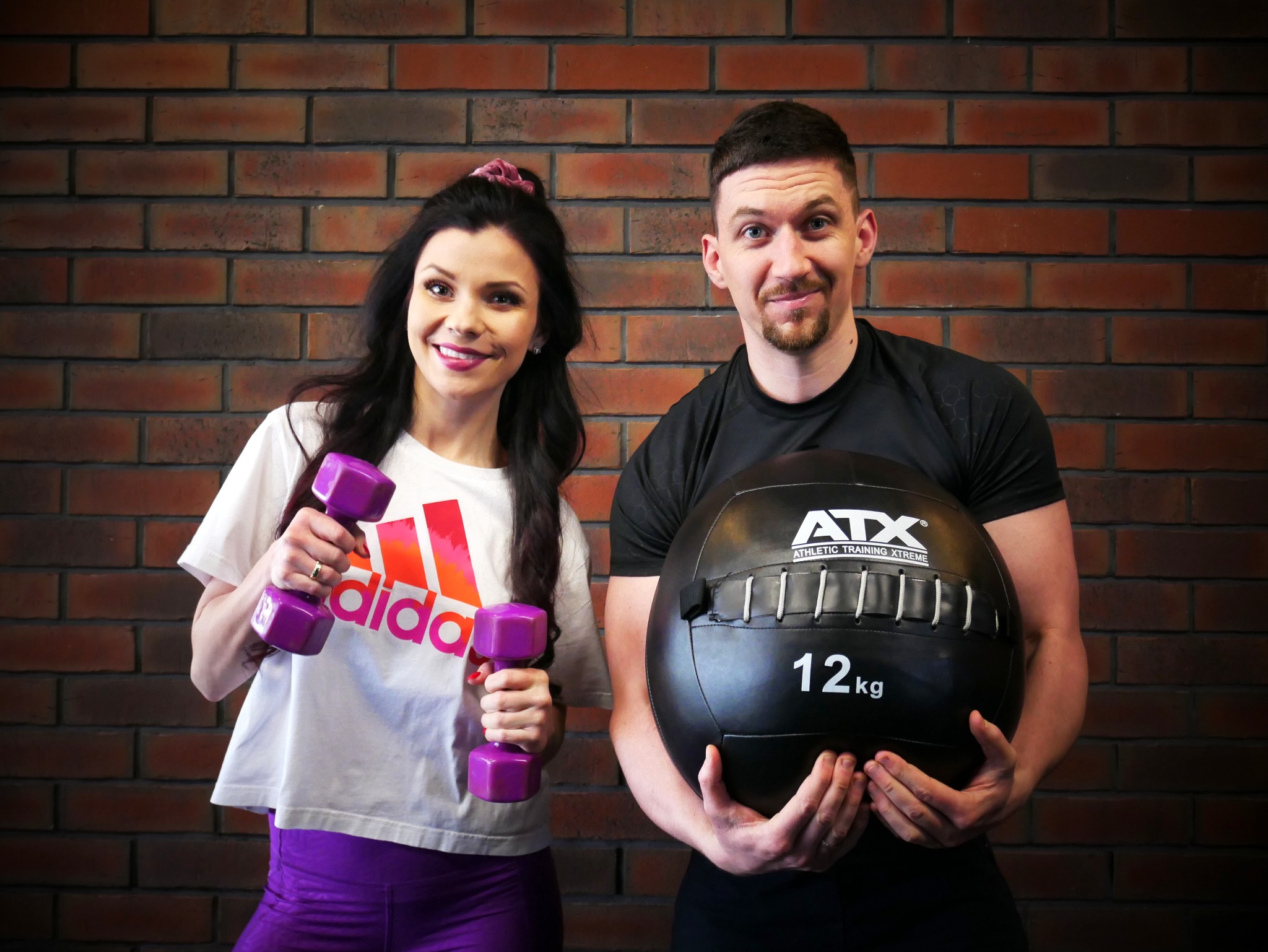 Uśmiechnięta kobieta trzymająca fioletowe hantle i mężczyzna trzymający czarną piłkę lekarską 12 kg na tle ściany z cegły.