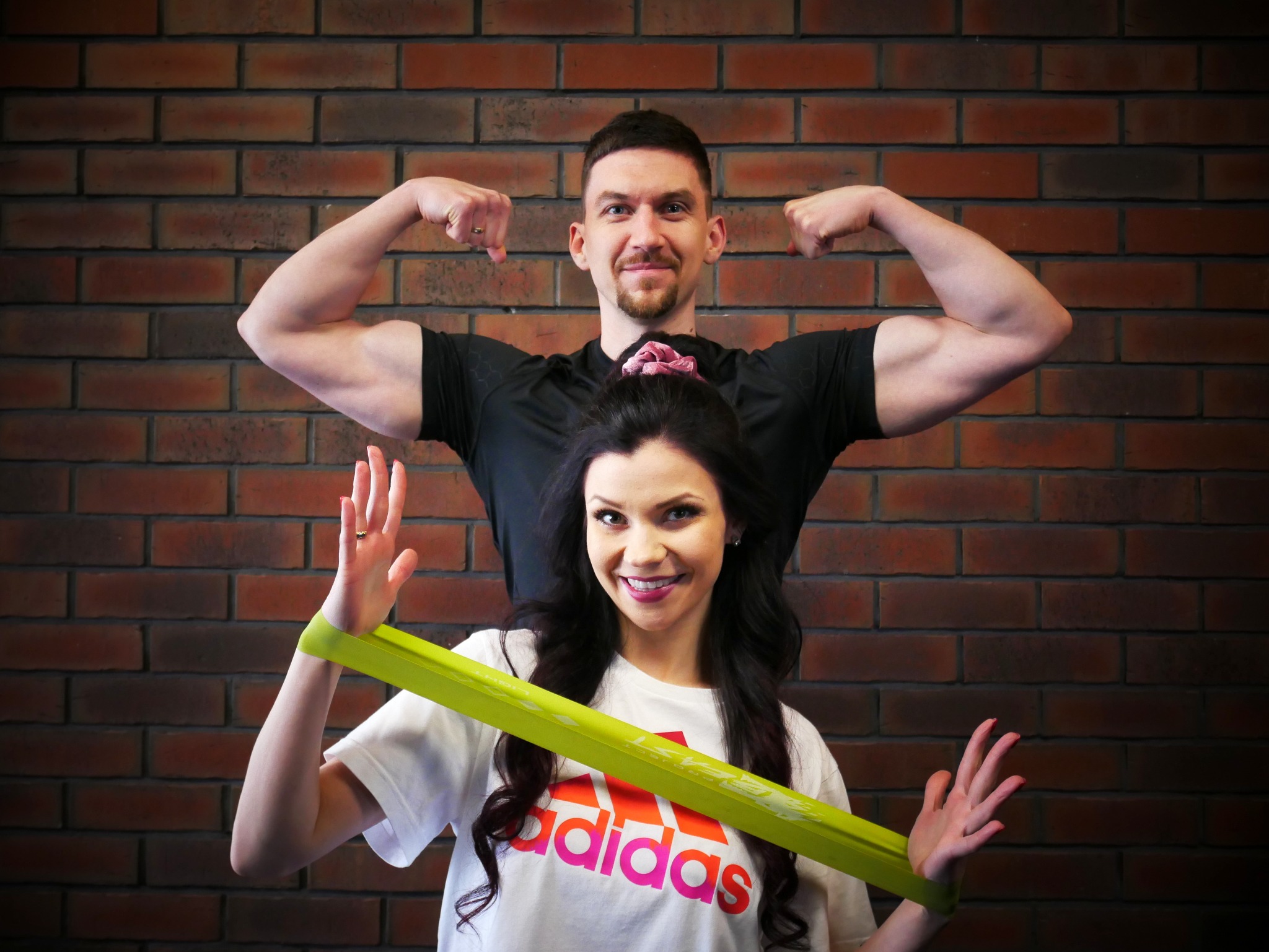 Uśmiechnięta kobieta z zieloną gumą do ćwiczeń na tle mężczyzny prezentującego bicepsy przed ceglaną ścianą.