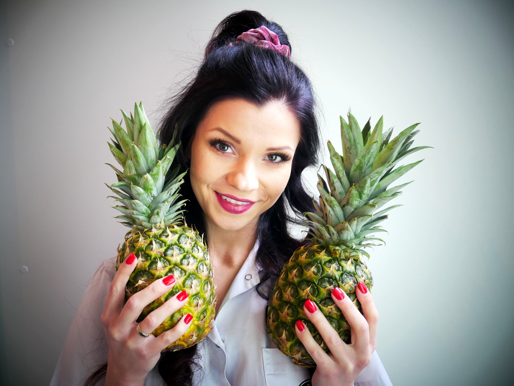 Uśmiechnięta kobieta w białej koszuli trzymająca dwa ananasy w dłoniach z czerwonym manicure na tle białej ściany.