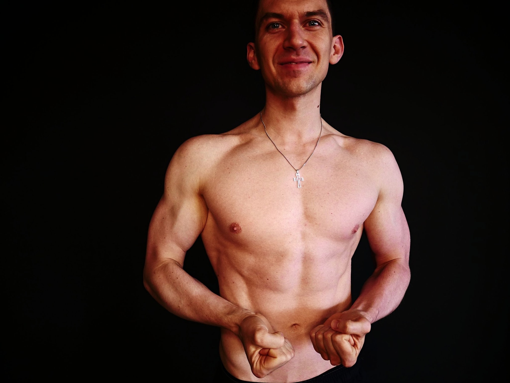 Uśmiechnięty mężczyzna z widocznymi mięśniami klatki piersiowej i bicepsami, noszący srebrny łańcuszek z krzyżykiem, na czarnym tle.