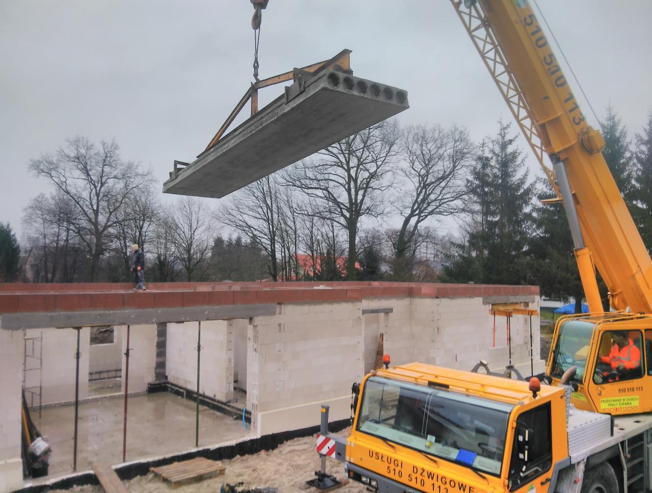 Żółty dźwig podnosi prefabrykowany betonowy strop na budowę domu z białych bloczków, widoczny pracownik na murze i drzewa w tle.