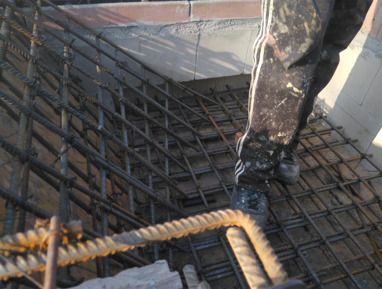 Zbrojenie pod fundamenty z widocznymi nogami robotnika w ubrudzonych spodniach dresowych i roboczych butach. Widoczne pręty zbrojeniowe tworzące siatkę, zalane częściowo betonem.
