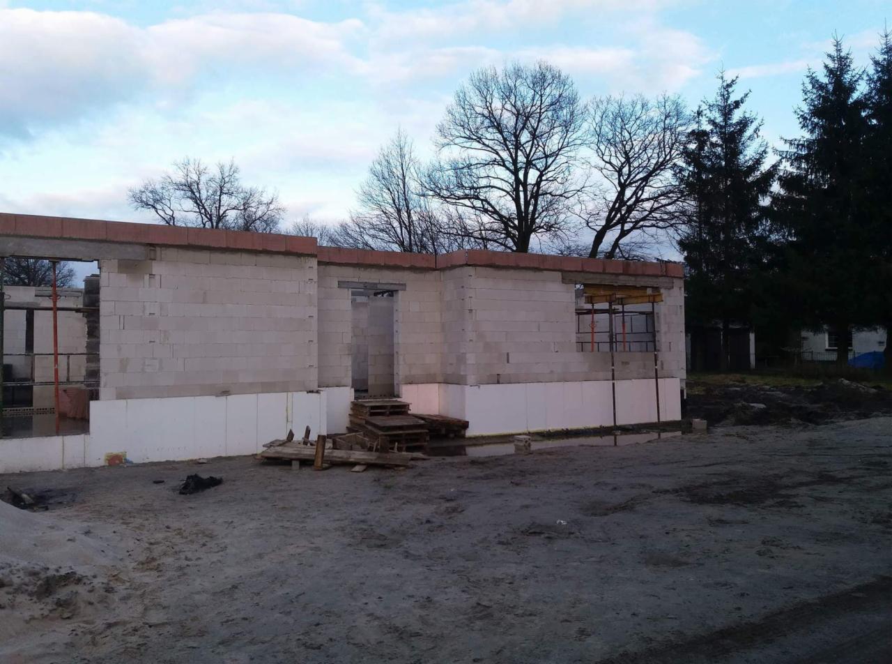 Wznoszenie ścian parterowego domu z betonu komórkowego na fundamencie z termoizolacją, widoczne zbrojenia i drewniane szalunki okienne oraz prowizoryczne schody z palet.