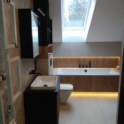 ADBUD Adam Bielawski - Nowoczesna łazienka na poddaszu z oknem dachowym, umywalką nablatową na czarnej szafce, podświetlaną wanną obudowaną drewnianymi panelami oraz grzejnikiem drabinkowym.