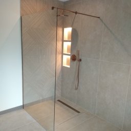 ADBUD Adam Bielawski - Nowoczesna kabina prysznicowa walk-in z szklaną ścianką, wykończona płytkami w odcieniach szarości, z dekoracyjną ścianką z geometrycznym wzorem i wnęką oświetloną od wewnątrz.