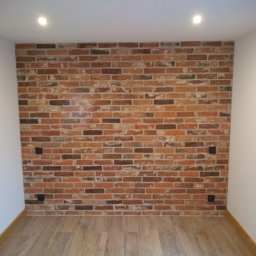 ADBUD Adam Bielawski - Ściana wykończona cegłą dekoracyjną w odcieniach brązu i czerwieni, z widocznymi puszkami elektrycznymi, podświetlona dwoma punktowymi lampami sufitowymi, z drewnianą podłogą w jasnym odcieniu.